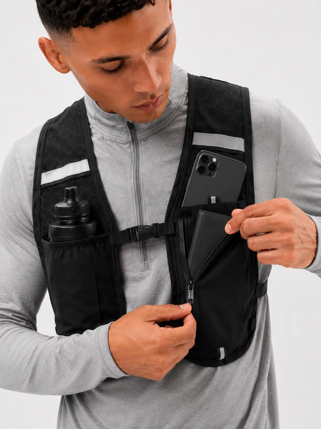 A man wearing a black running chest harness vest over a gray long-sleeve top, shown from the chest up. He is demonstrating the vest's pockets, with one side holding a black water bottle and the other holding a black smartphone in a zippered pocket.