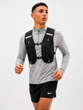 A man wearing a black running hydration backpack vest over a gray long-sleeve athletic shirt and black Nike running shorts, captured against a plain light gray studio background.