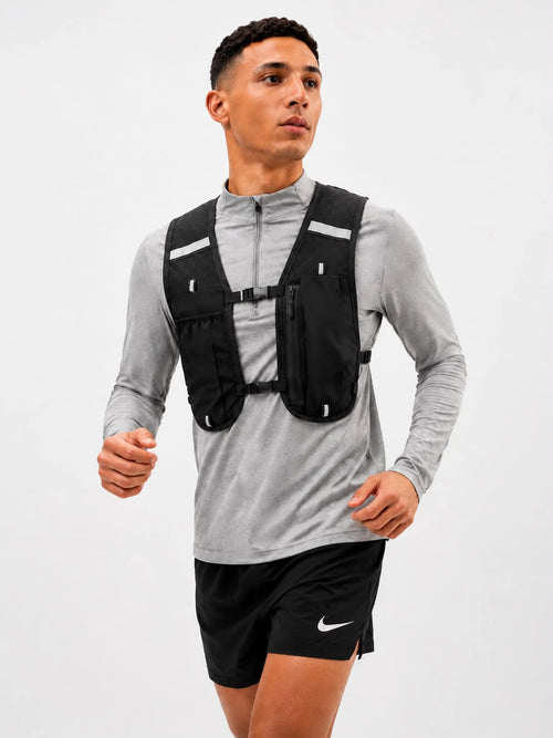A man wearing a black running hydration backpack vest over a gray long-sleeve athletic shirt and black Nike running shorts, captured against a plain light gray studio background.