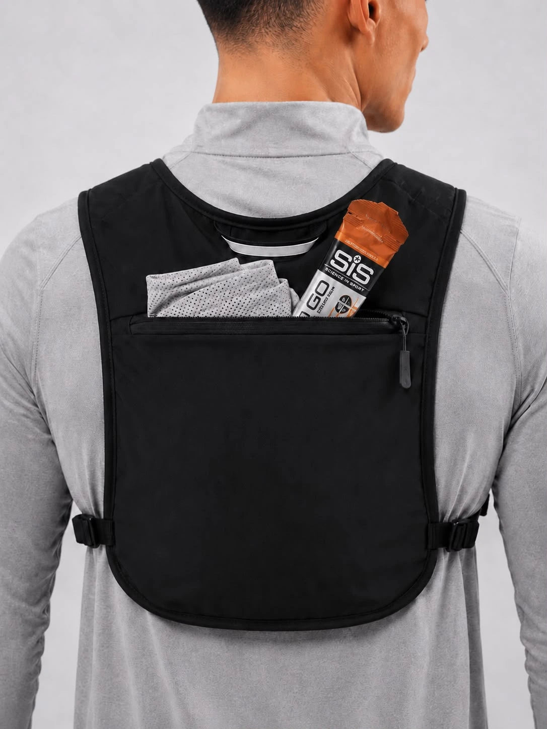A man viewed from behind wearing a black athletic backpacking vest over a light gray long-sleeve shirt. The vest has a front zippered pocket containing a gray towel and a protein bar, with elastic side straps and a zippered neck opening.
